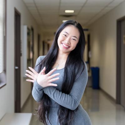 A smiling young woman with long black hair stands in a hallway with her arms crossed. She exudes confidence and warmth, representing the welcoming environment of the AAPI center.